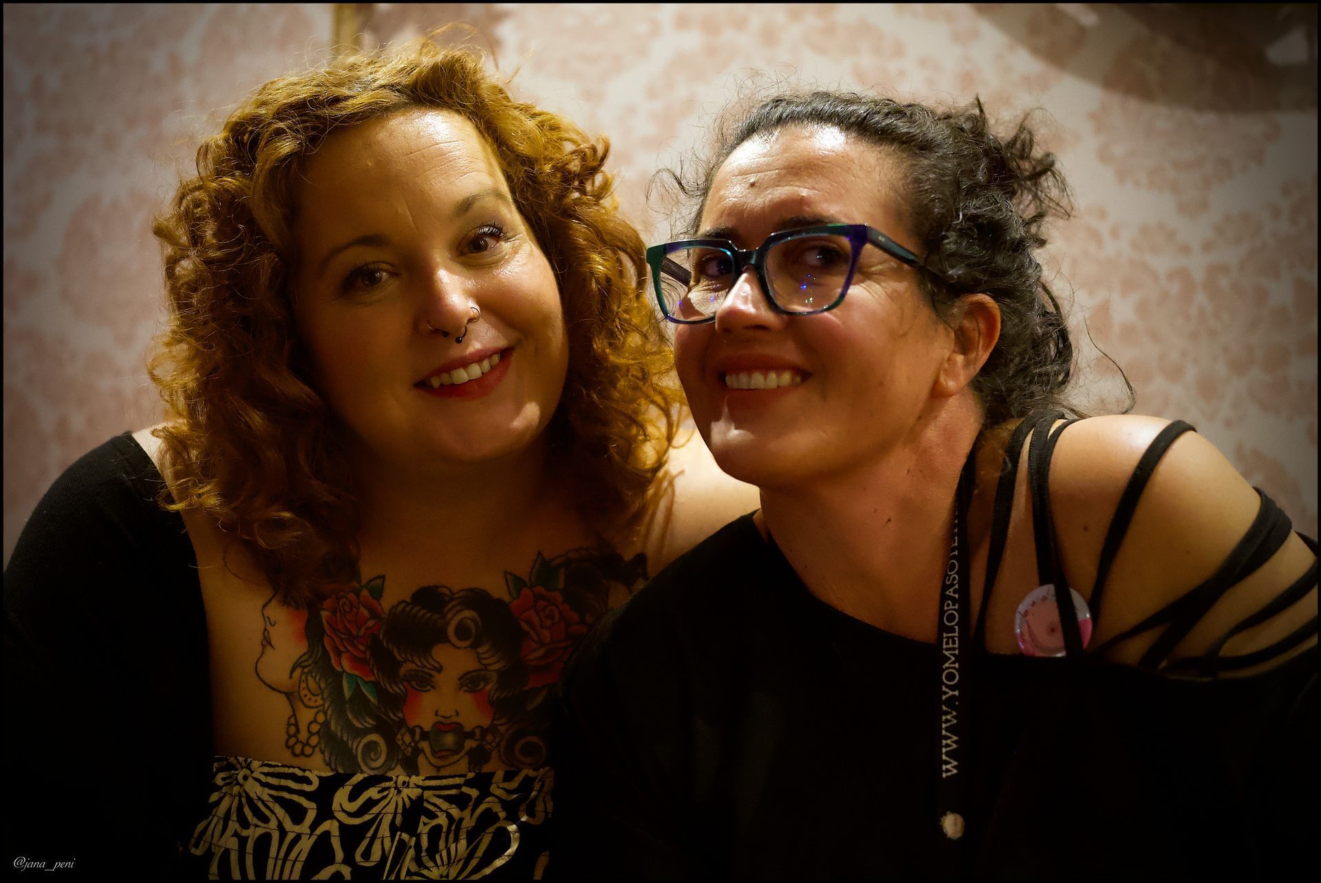 Dos mujeres sonrientes posan juntas. Una con cabello rizado y un tatuaje, la otra con gafas y cabello oscuro, frente a una pared estampada.