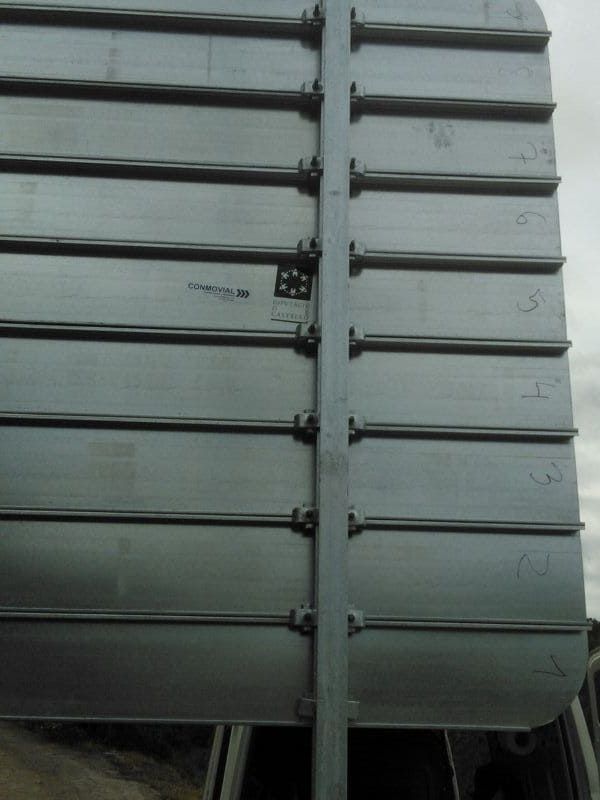 Parte posterior de un letrero metálico con listones horizontales, fijado a un poste vertical con herrajes.