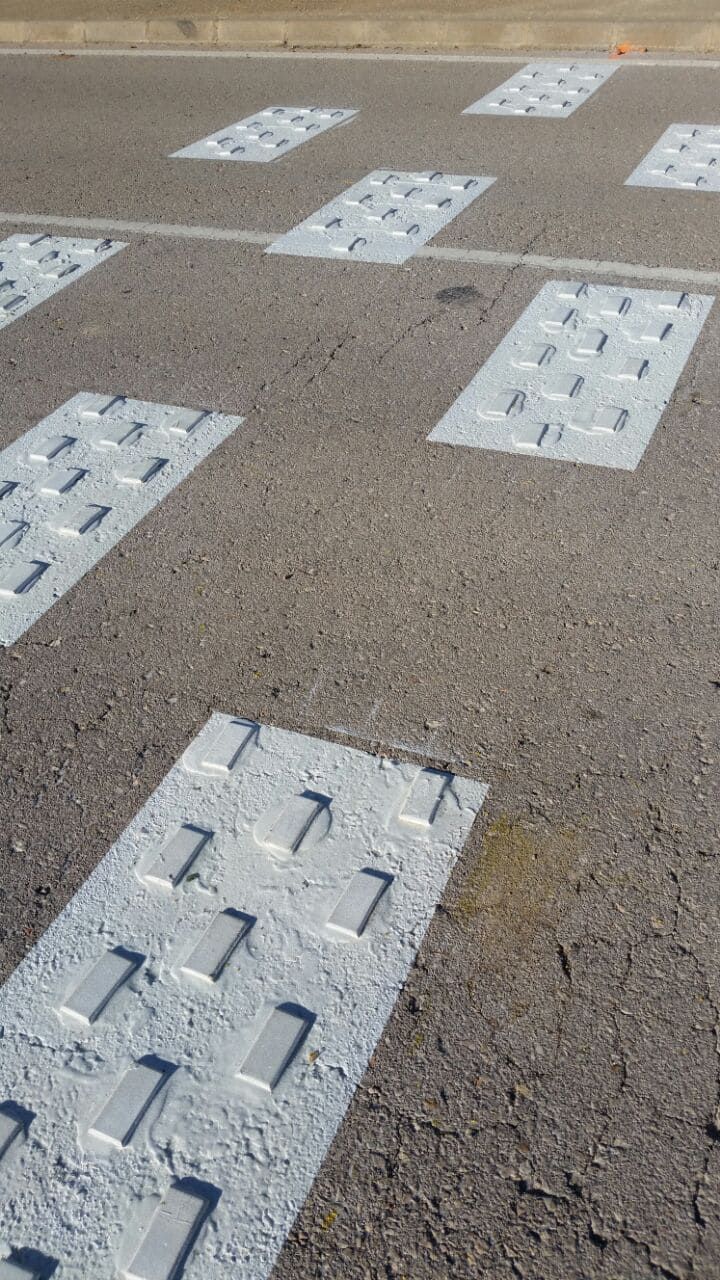 Carretera asfaltada con parches rectangulares pintados. Cada parche tiene formas rectangulares en relieve, táctiles y aptas para personas con discapacidad visual.