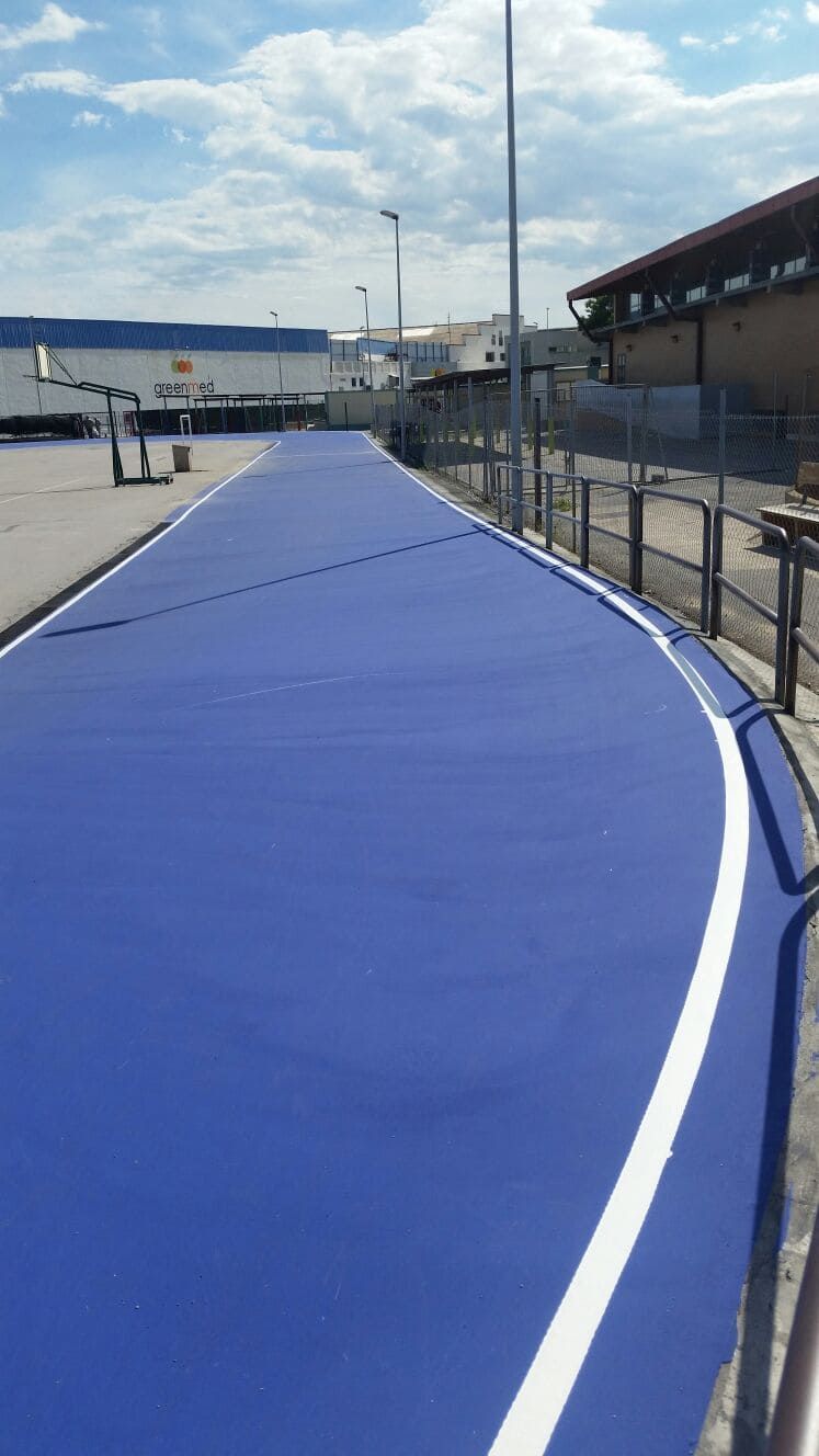 Pasarela azul con lineas blancas y barandilla metálica, junto a un edificio y zona de aparcamiento con un camión.