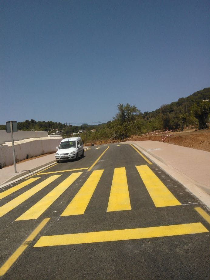 Furgoneta blanca acercándose a un cruce peatonal con rayas amarillas y negras en una carretera pavimentada, con montañas y cielo azul de fondo.