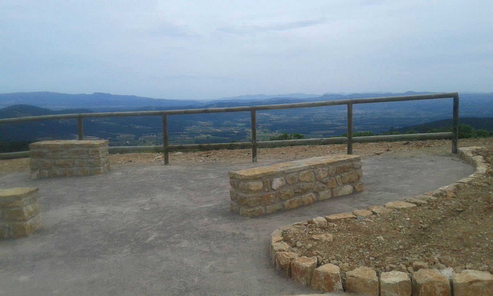 Mirador con bancos de piedra, barandilla de madera y vista de montañas en capas bajo un cielo nublado.