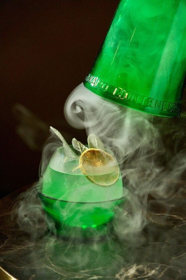 Botella y vaso de color verde con humo arremolinado, iluminados con luz verde neón, con una rodaja de limón.