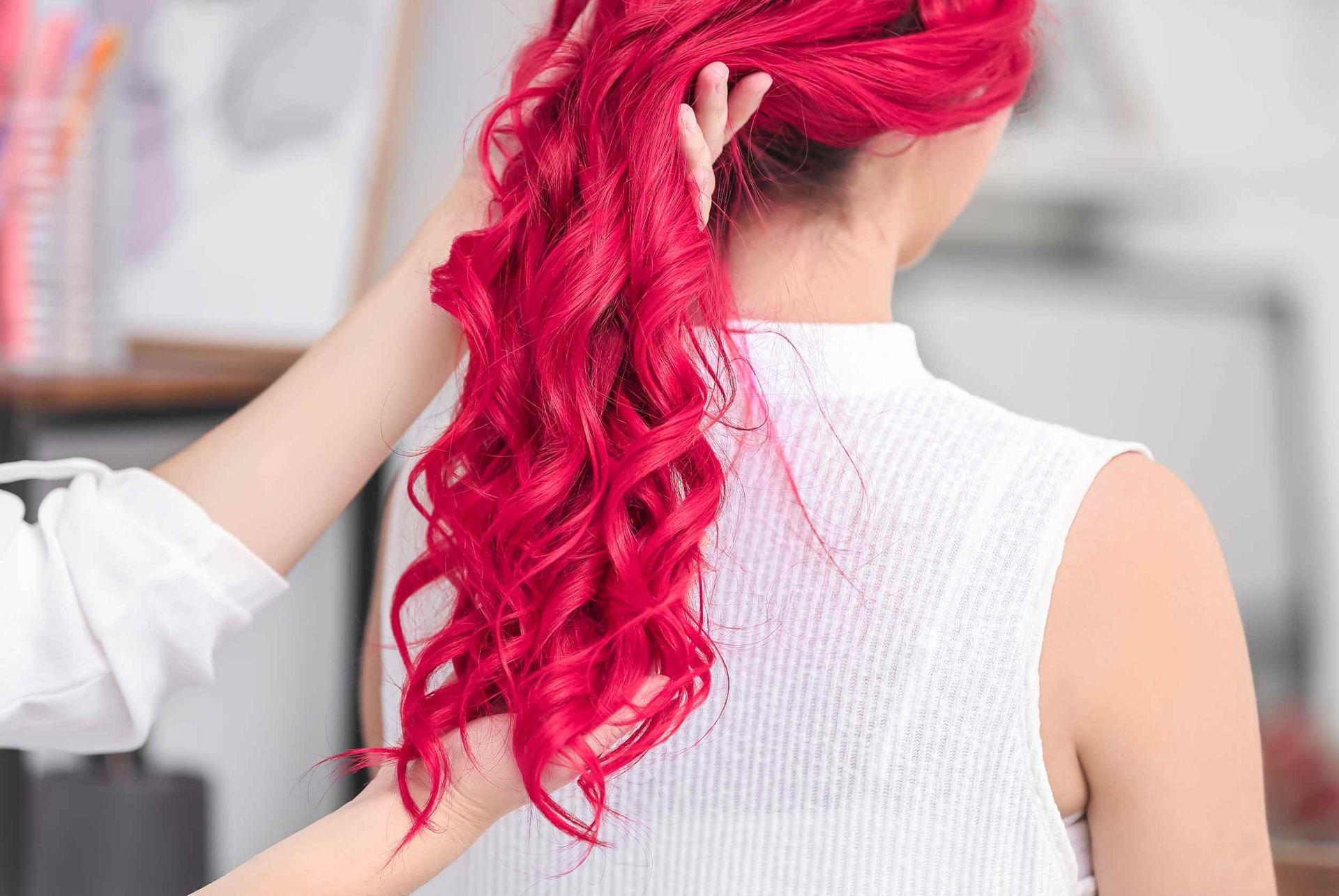 Mujer con cabello rizado rojo brillante, vista de espaldas. Una mano arreglándose el cabello. Blusa blanca, luz natural.