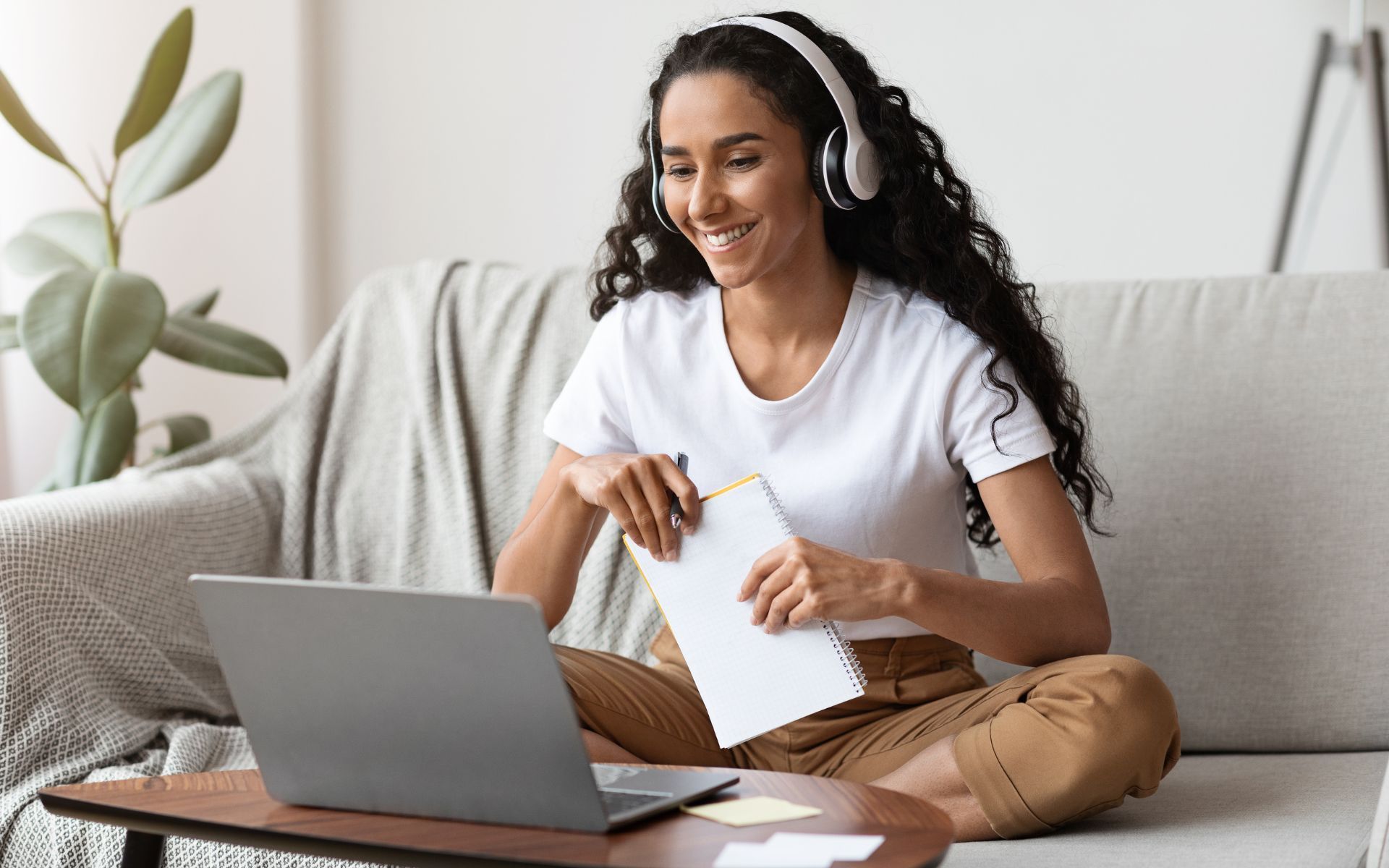 Mujer con auriculares, computadora portátil y bloc de notas sonríe en un sofá.