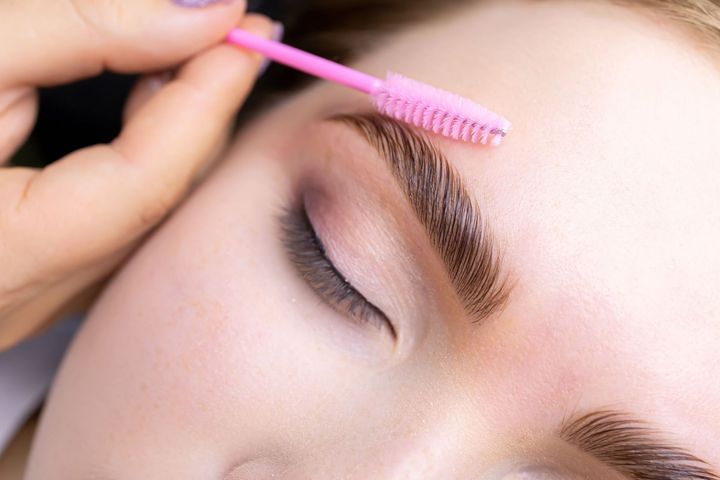 Cejas de una persona siendo depiladas con un cepillo rosa. Primer plano del rostro, resaltando las cejas y el maquillaje de ojos.
