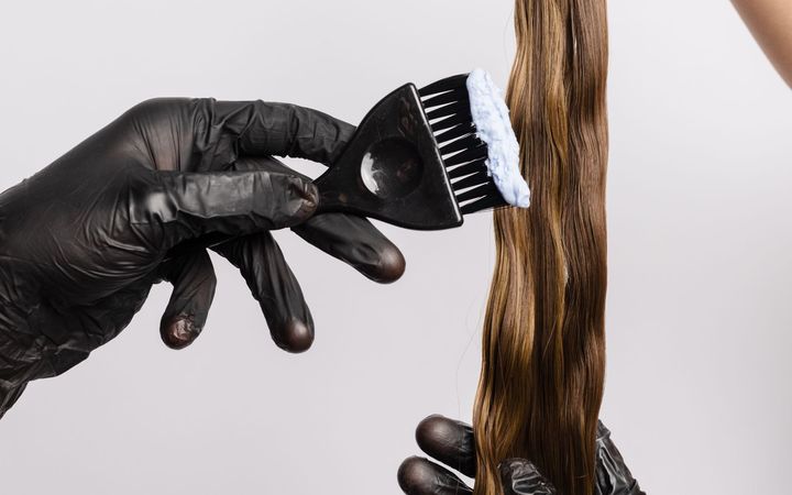 Persona que usa guantes negros aplica tinte para el cabello en mechones de cabello castaño con un cepillo negro.