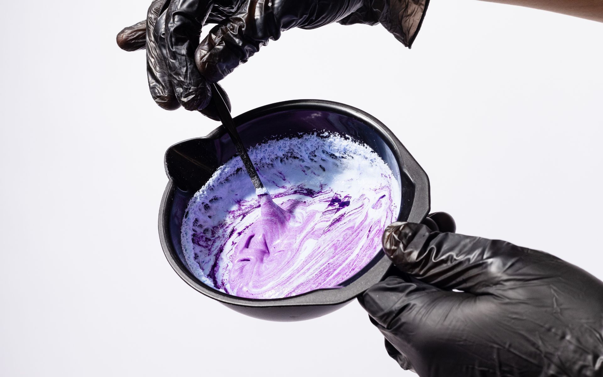Manos con guantes negros mezclando tinte para el cabello morado en un recipiente.