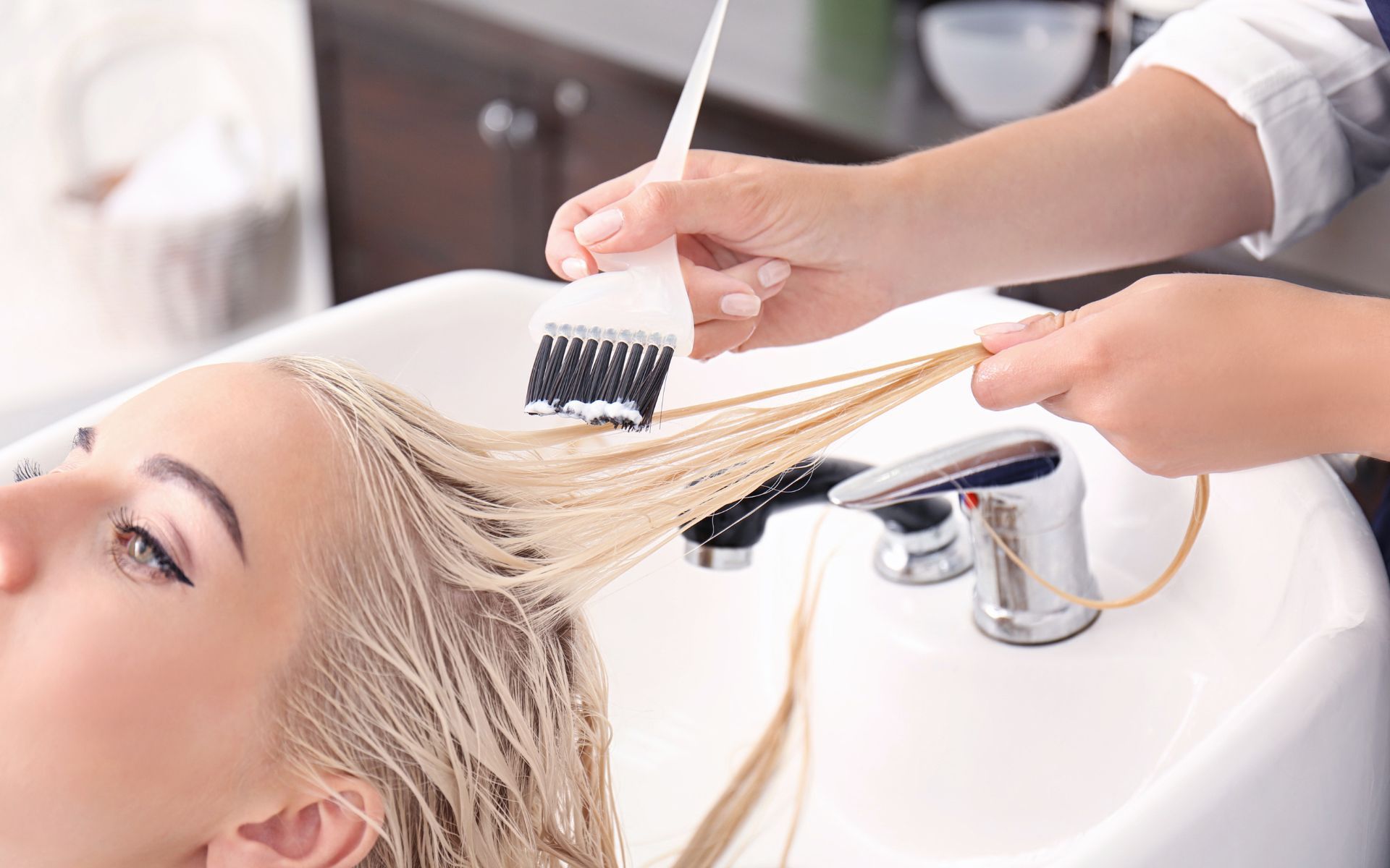 Peluquera aplicando tinte para el cabello al cabello rubio de una mujer en el lavabo de un salón.