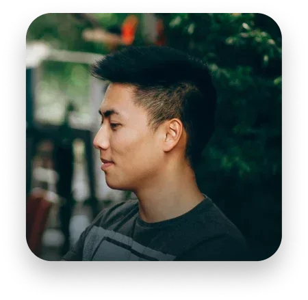 Man in profile, dark hair, wearing a gray shirt, outside with blurred greenery in the background.