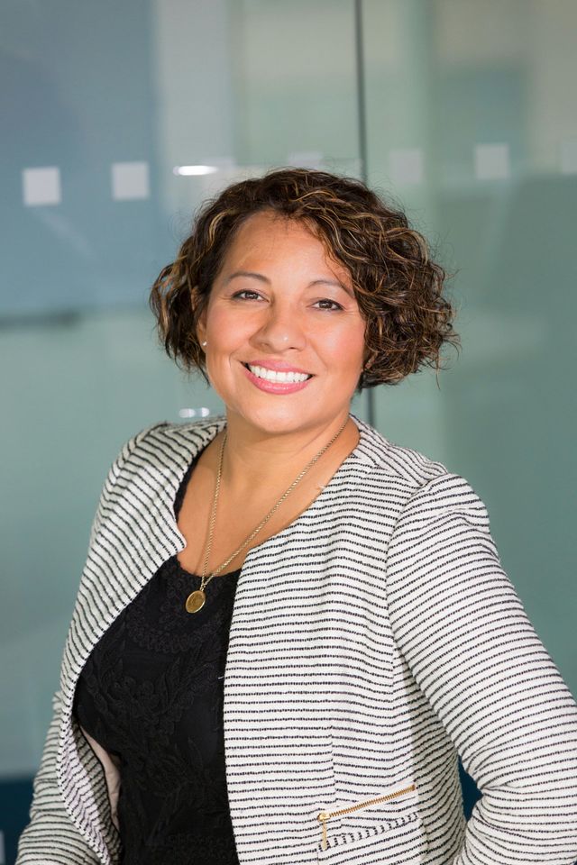 Woman smiling, wearing a black top and striped jacket, near a glass wall.