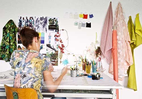 Eine Frau sitzt an einem Schreibtisch vor einer Wand, an der Kleidung hängt.