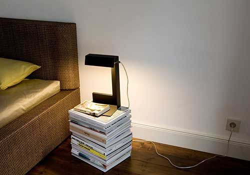 Auf einem Stapel Bücher neben einem Bett steht eine Lampe.