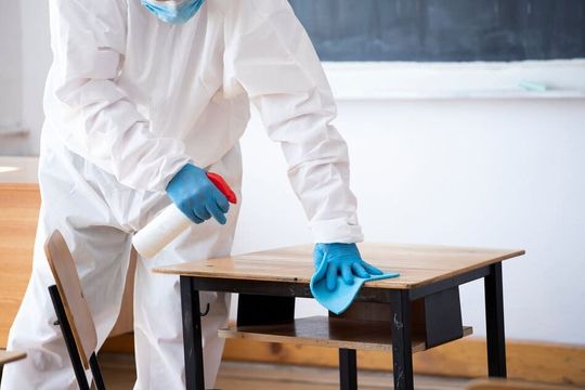 Una persona con un traje de protección está limpiando un escritorio en un aula.