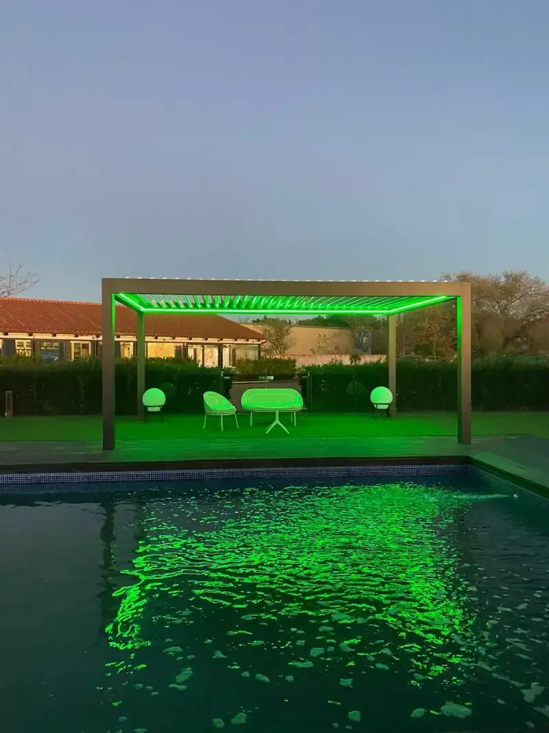 Una pérgola está iluminada con luces verdes junto a una piscina.