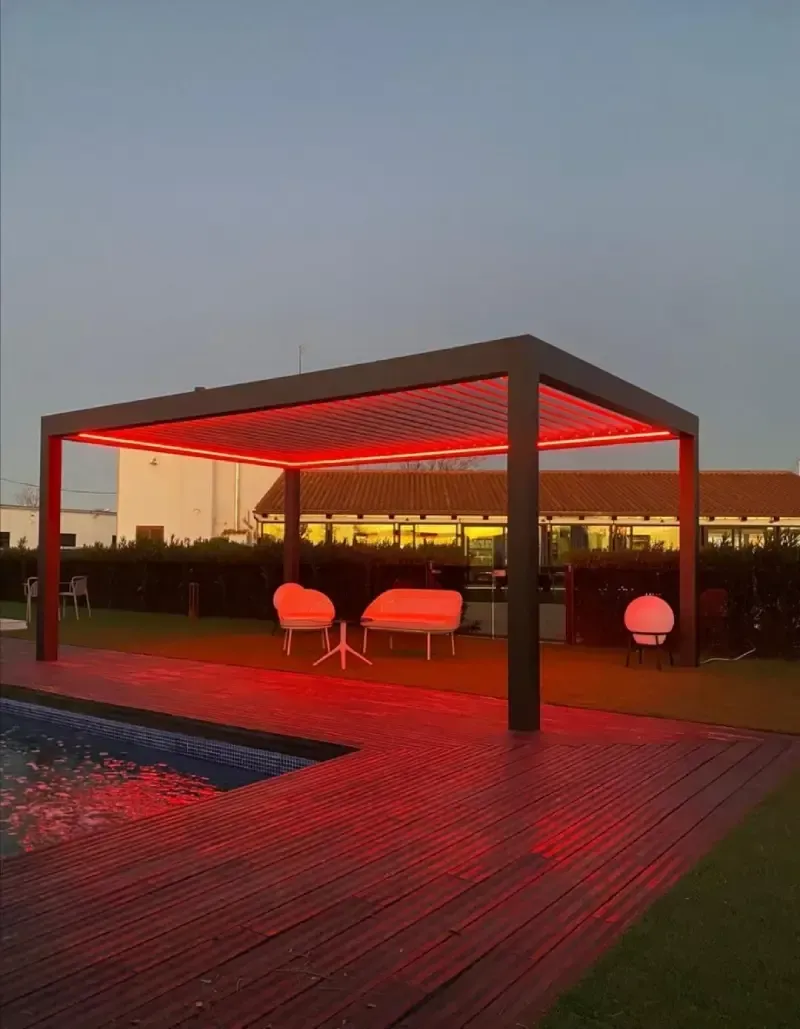 Una pérgola está iluminada con luces rojas junto a una piscina.