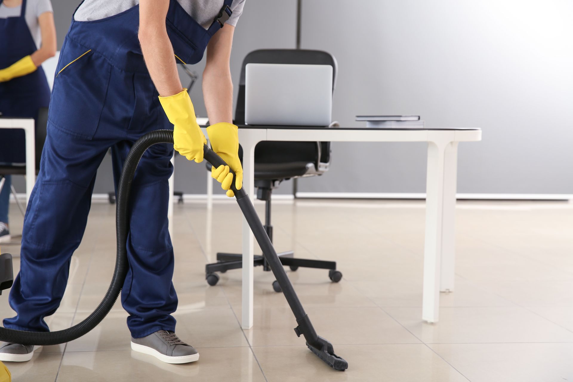 Agent d'entretien passant l'aspirateur sur le sol d'un bureau à côté d'une chaise, portant des gants jaunes et un uniforme bleu