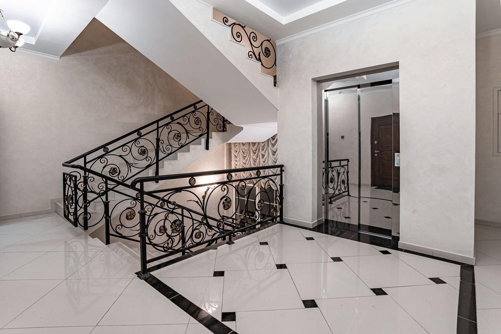Escalier intérieur avec rampes noires, sol carrelé blanc et porte d'ascenseur à miroir.