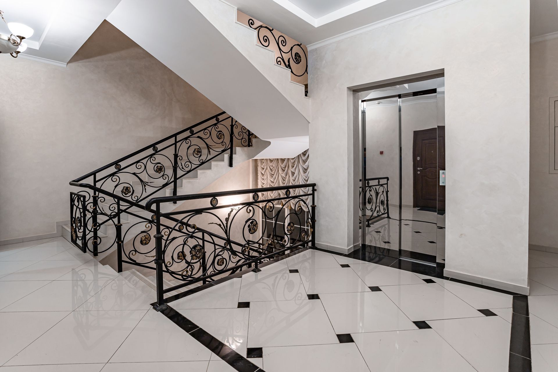 Élégant escalier intérieur avec rampes en fer forgé noir, sol carrelé blanc et porte d'ascenseur.