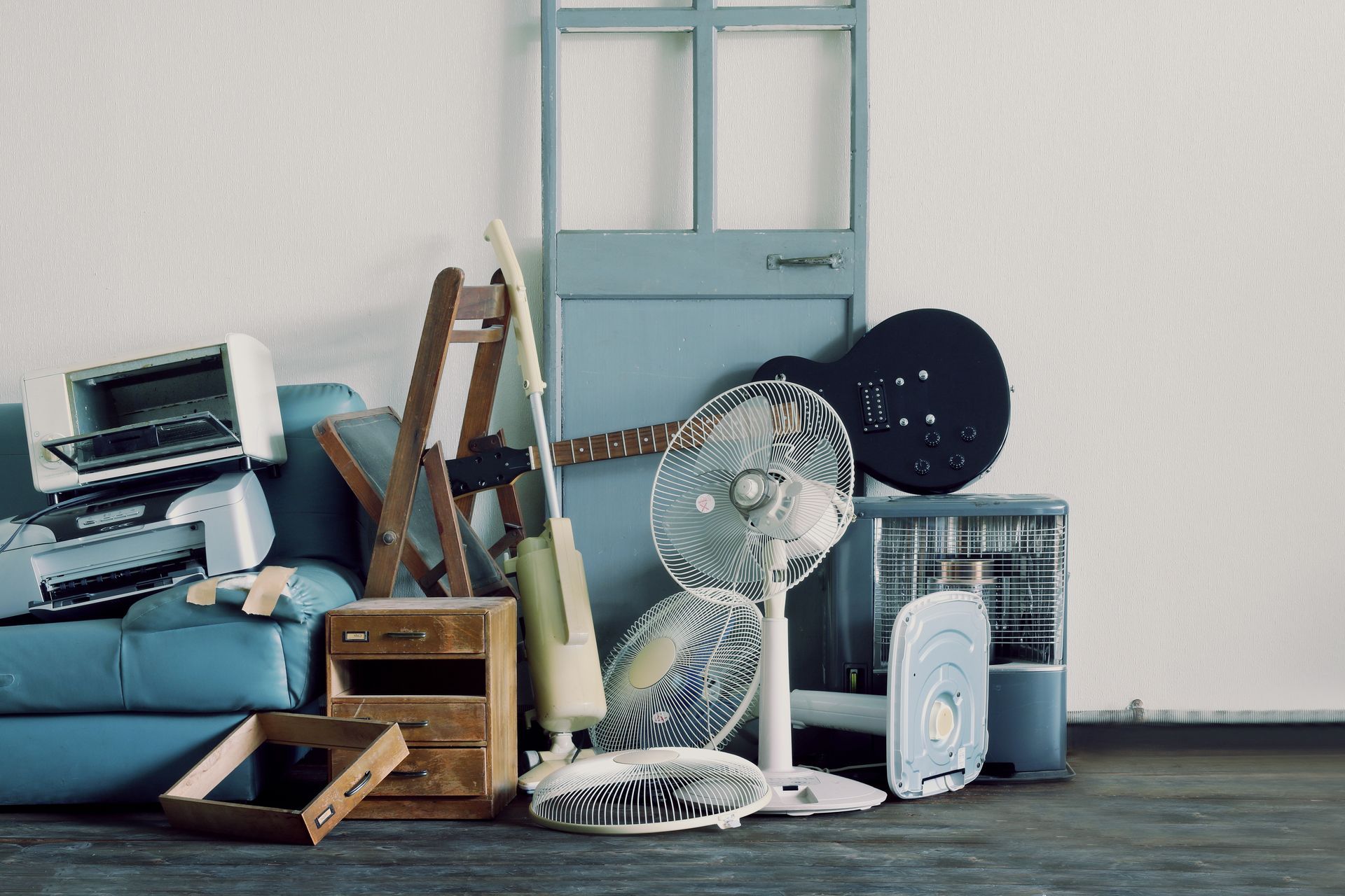 Coin de pièce encombré avec des cartons empilés, un ventilateur et des objets ménagers appuyés contre une porte bleue.