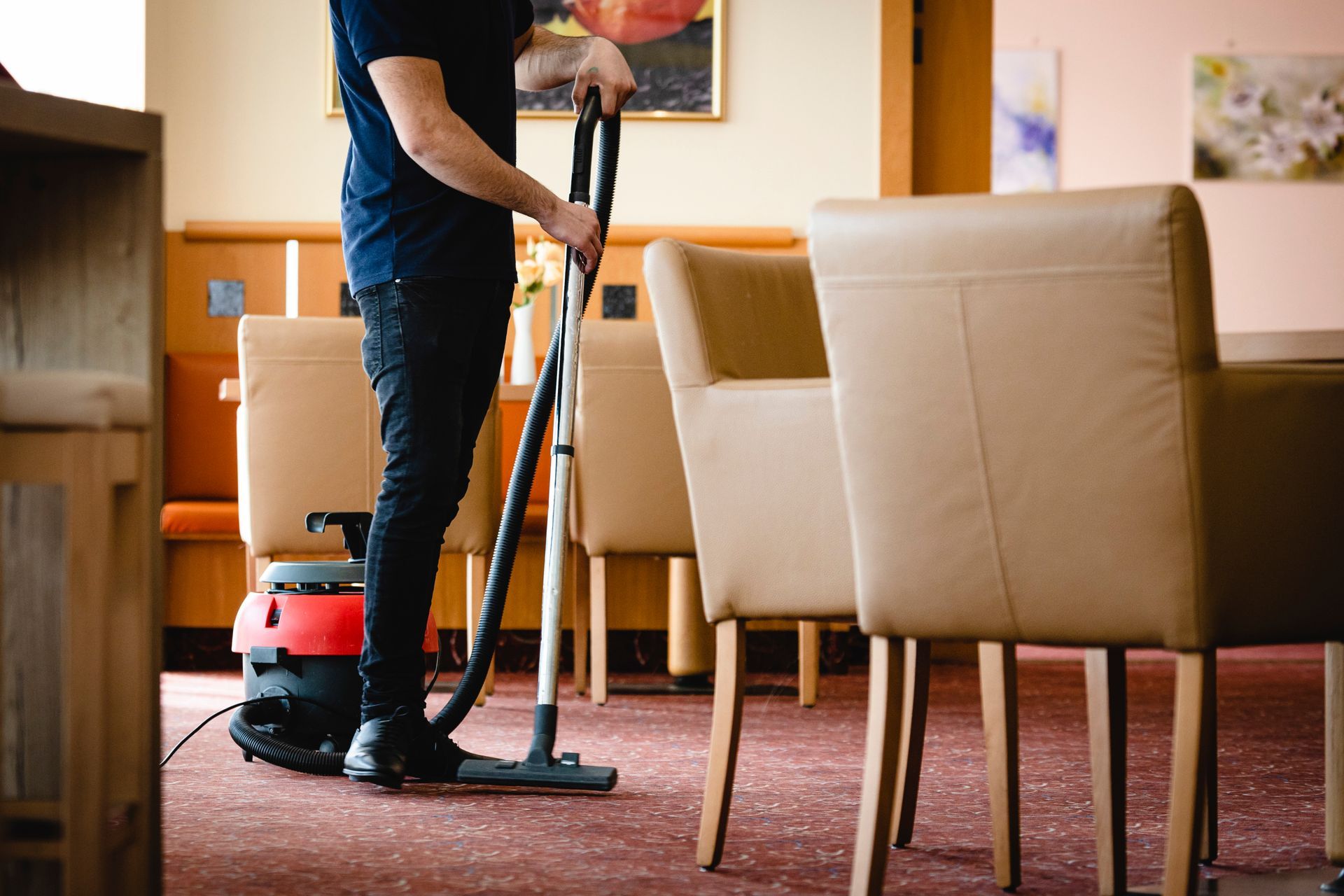 Une personne passe l'aspirateur dans une pièce recouverte de moquette avec un aspirateur traîneau rouge, à côté de chaises beiges.