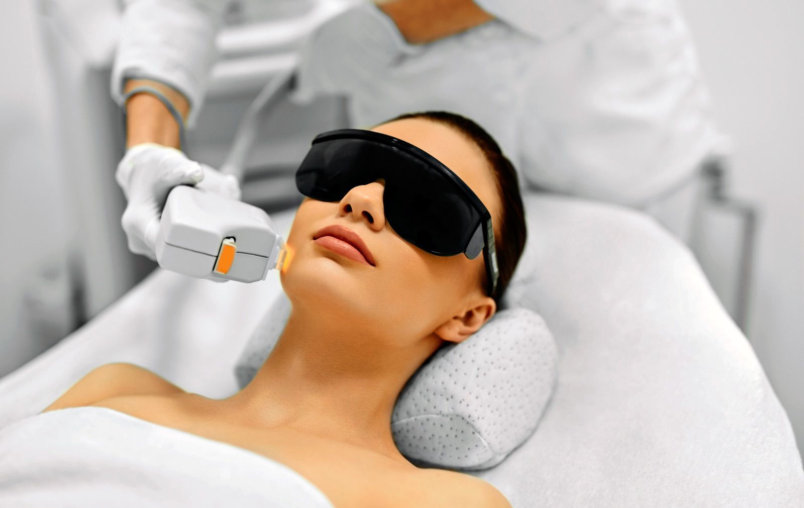 Mujer recibe un tratamiento láser facial mientras usa gafas protectoras, acostada en una cama médica en una habitación blanca.