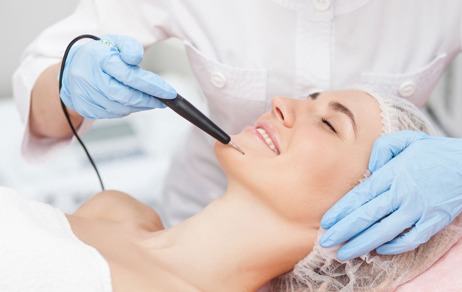 Una persona recibe un tratamiento cosmético facial con un dispositivo portátil. El profesional usa guantes y sujeta el rostro de la persona.