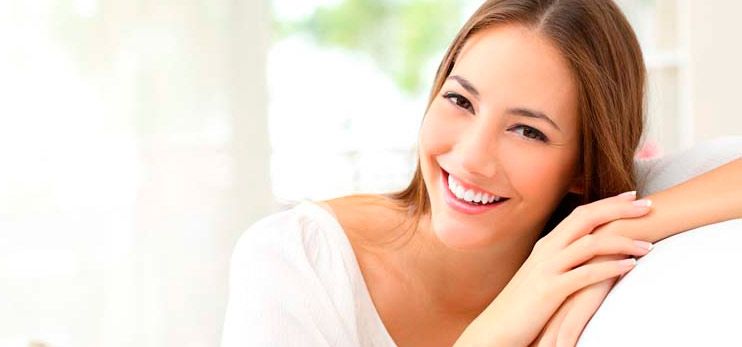 Una mujer de cabello castaño y sonrisa brillante está sentada en el interior, apoyando su brazo sobre una superficie blanca, con un fondo borroso.