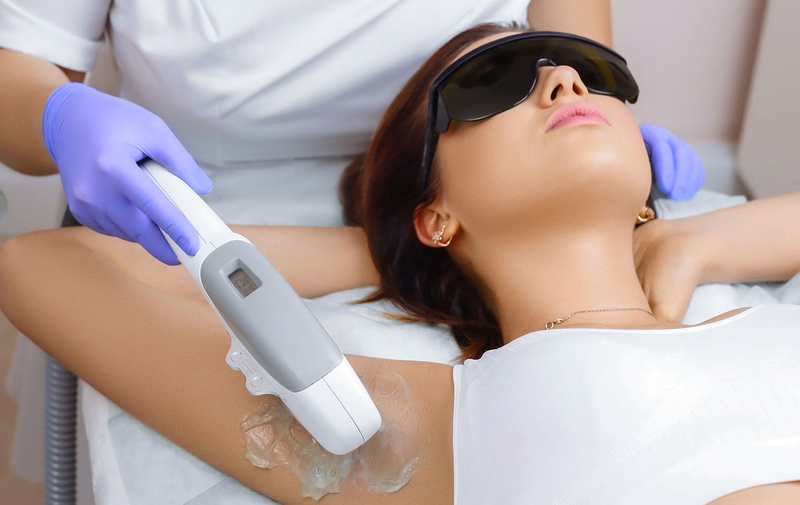 Una mujer recibe depilación láser en la axila en una clínica. Un técnico utiliza un dispositivo láser mientras la mujer usa gafas protectoras.