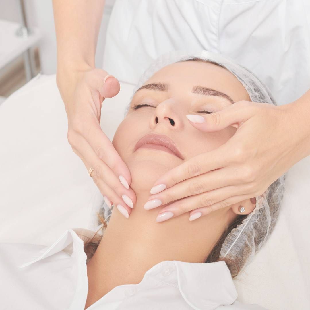 Una mujer recibe un masaje facial, tumbada con los ojos cerrados y las manos sobre el rostro. Lleva una redecilla y ropa blanca en una sala de tratamiento.
