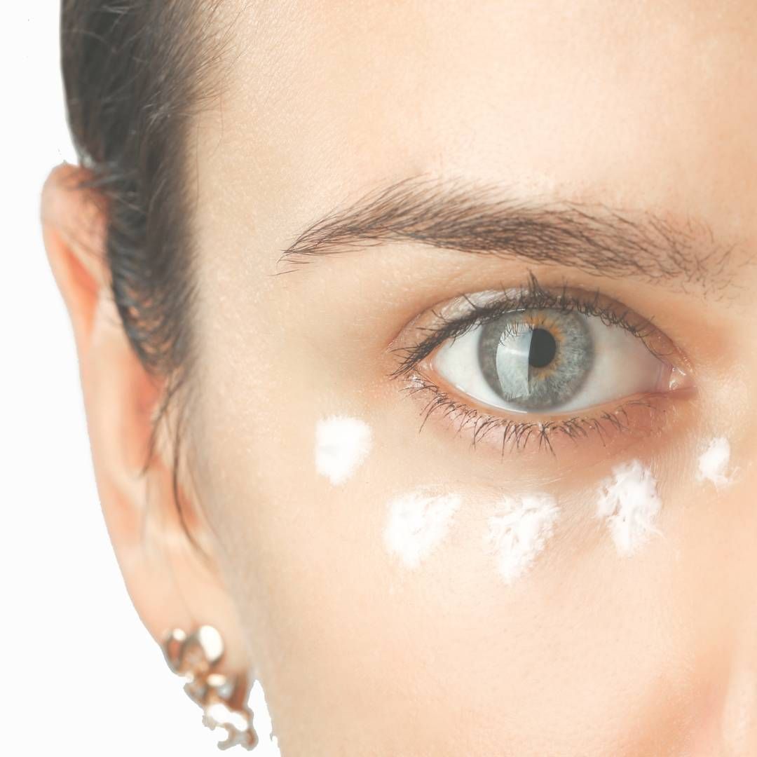 Primer plano del ojo de una mujer con puntos de crema aplicados debajo, probablemente para el cuidado de la piel. Ojo azul, piel pálida, expresión neutra.