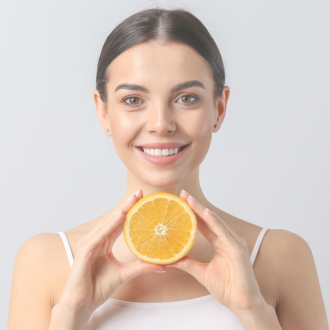 Mujer sonriendo con una rodaja de naranja. Lleva una camiseta blanca sin mangas sobre un fondo liso.