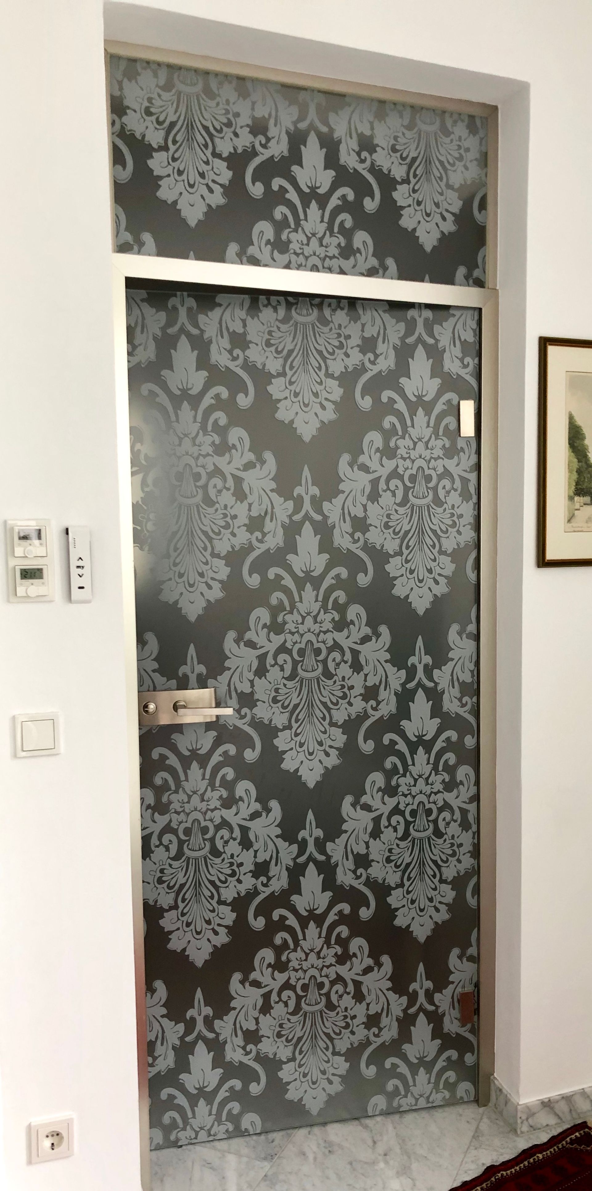 Glastür mit Damastmuster und silberfarbenen Beschlägen. Die Tür ist in eine weiße Wand eingelassen.