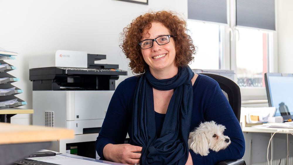 Eine Frau sitzt mit einem Hund auf ihrem Schoß an einem Schreibtisch.