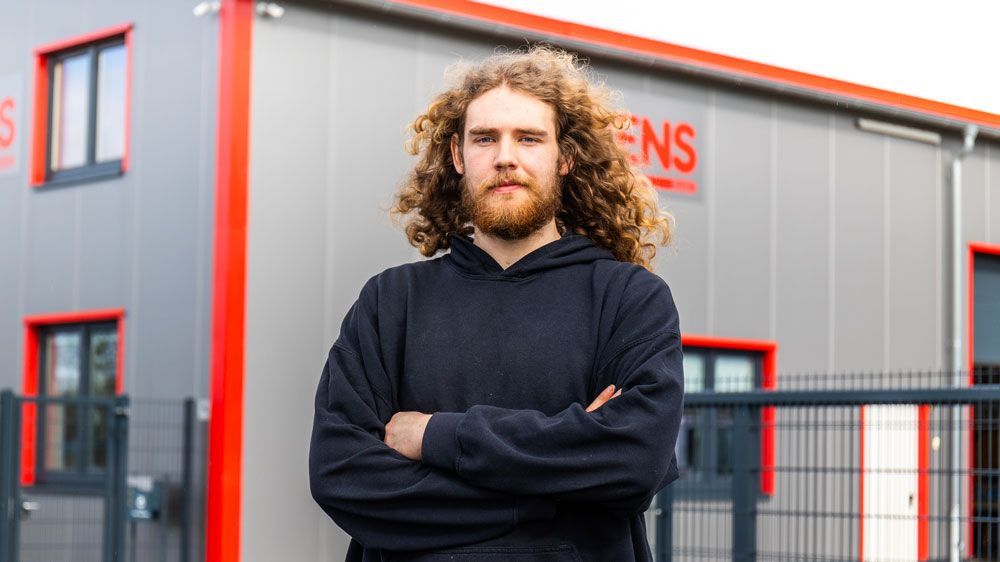 Ein Mann mit lockigem Haar und Bart steht mit verschränkten Armen vor einem Gebäude.