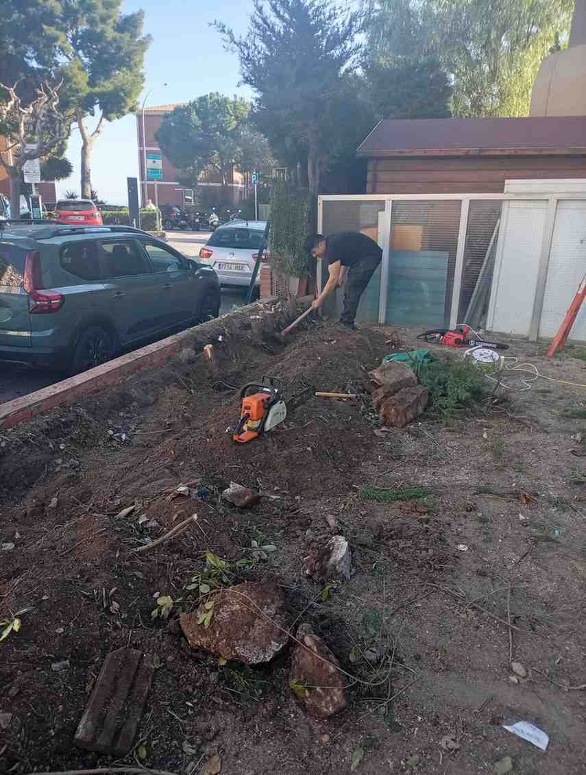 Hombre cavando en un cantero de jardín con una motosierra cerca, al lado de un coche y un edificio.