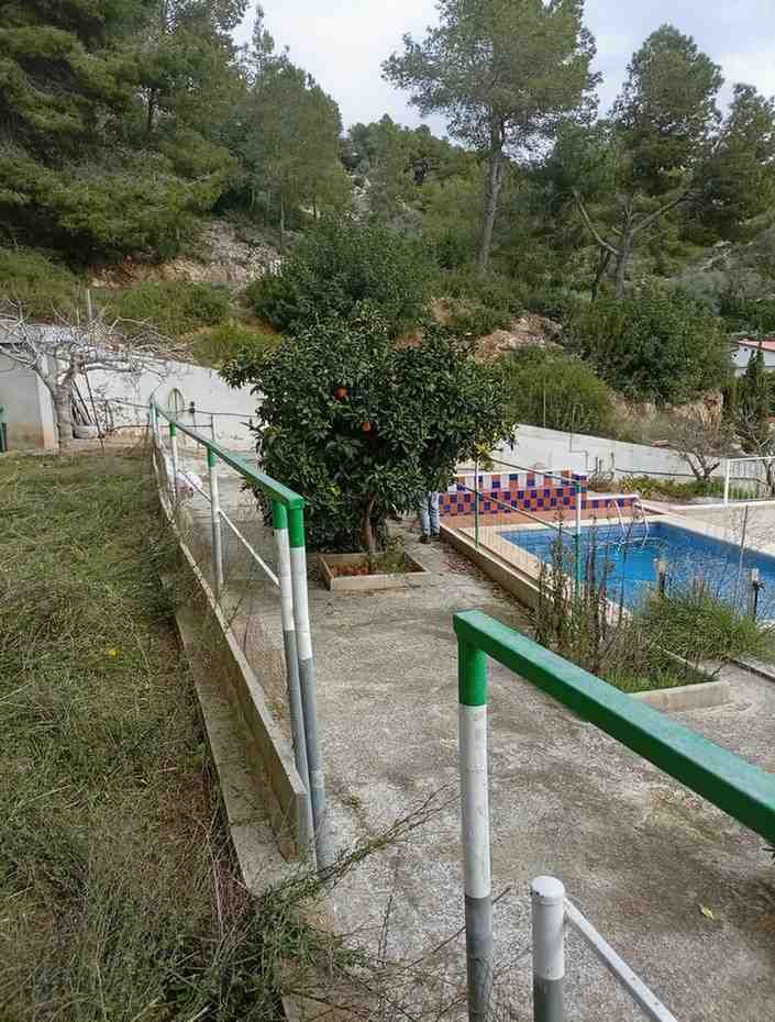 Camino de hormigón con barandilla verde, que conduce a una zona de piscina con árboles en una ladera.