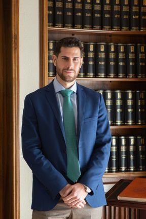 Hombre con traje azul y corbata verde, de pie frente a una estantería llena de libros.