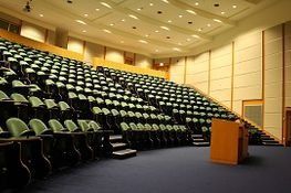 Auditorio con gradas, alfombra azul y podio. Paredes y techo beige con iluminación empotrada.