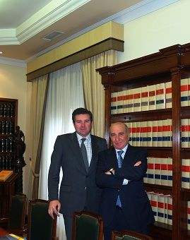 Hombre de traje sentado en un escritorio de oficina. El escritorio tiene lámpara, teléfono y papeles.