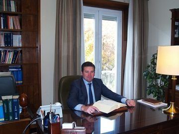 Hombre de traje sentado en un escritorio, mirando un libro grande en una oficina bien iluminada con estanterías y una ventana.