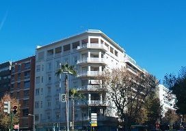 Edificio blanco de varios pisos con balcones, árboles y señales de calle bajo un cielo azul.