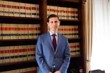 Un hombre con traje azul y corbata violeta se encuentra frente a una estantería llena de libros.