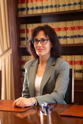 Una mujer con chaqueta y gafas está sentada en un escritorio frente a una estantería.