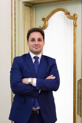 Un hombre con un traje azul y los brazos cruzados se encuentra junto a una puerta decorativa en una habitación con molduras ornamentadas.