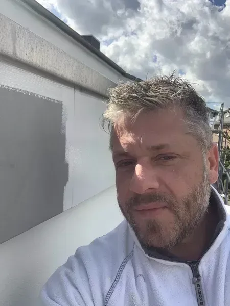 Ein Mann mit Bart steht vor einer weißen Wand.