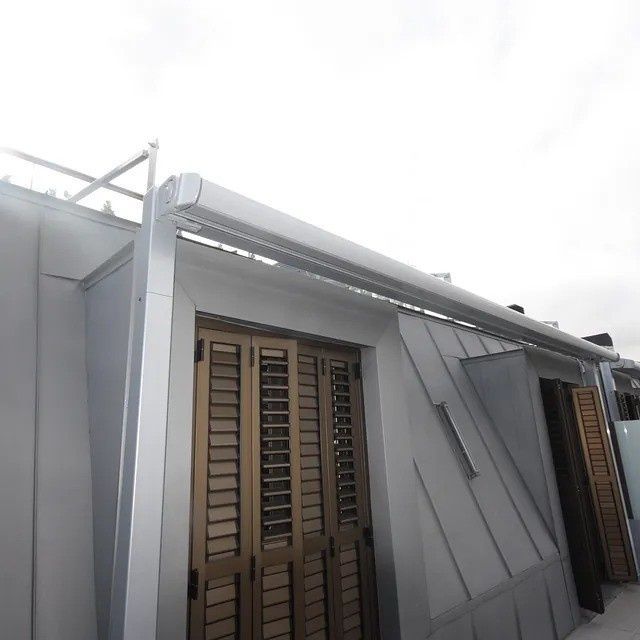 Exterior de una casa gris con puertas de celosía marrones y techo inclinado bajo un cielo nublado.