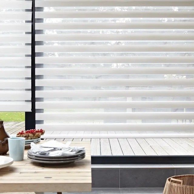 Patio moderno con persianas blancas, mesa de madera y silla de mimbre junto a una ventana con tarima de madera.