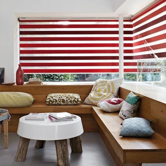 Acogedor rincón con persianas de rayas rojas y blancas, banco de madera, cojines y una pequeña mesa redonda.