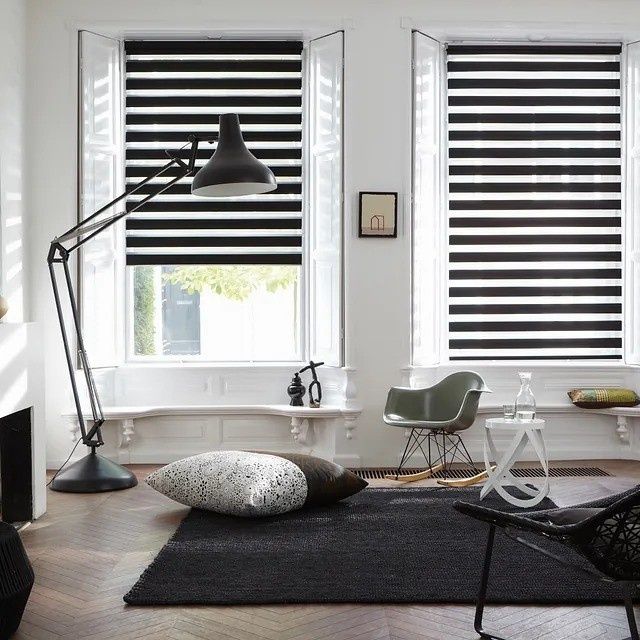 Sala de estar moderna con persianas blancas y negras, lámpara de pie, cojines estampados y alfombra oscura.